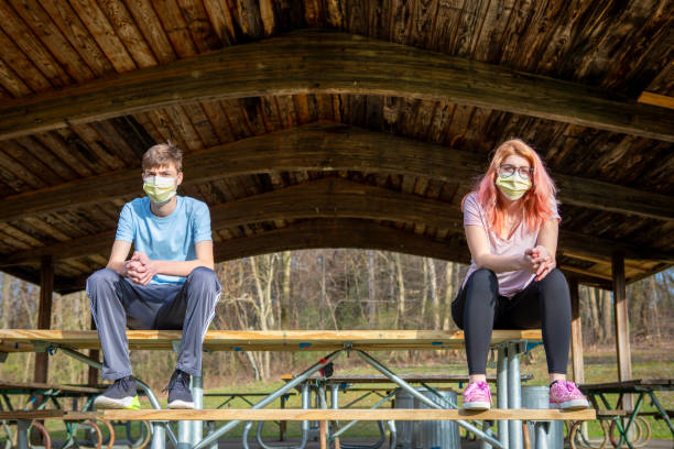 Teenage siblings wearing face masks sitting at a park grove outdoors keeping a social distance during the coronavirus pandemic.