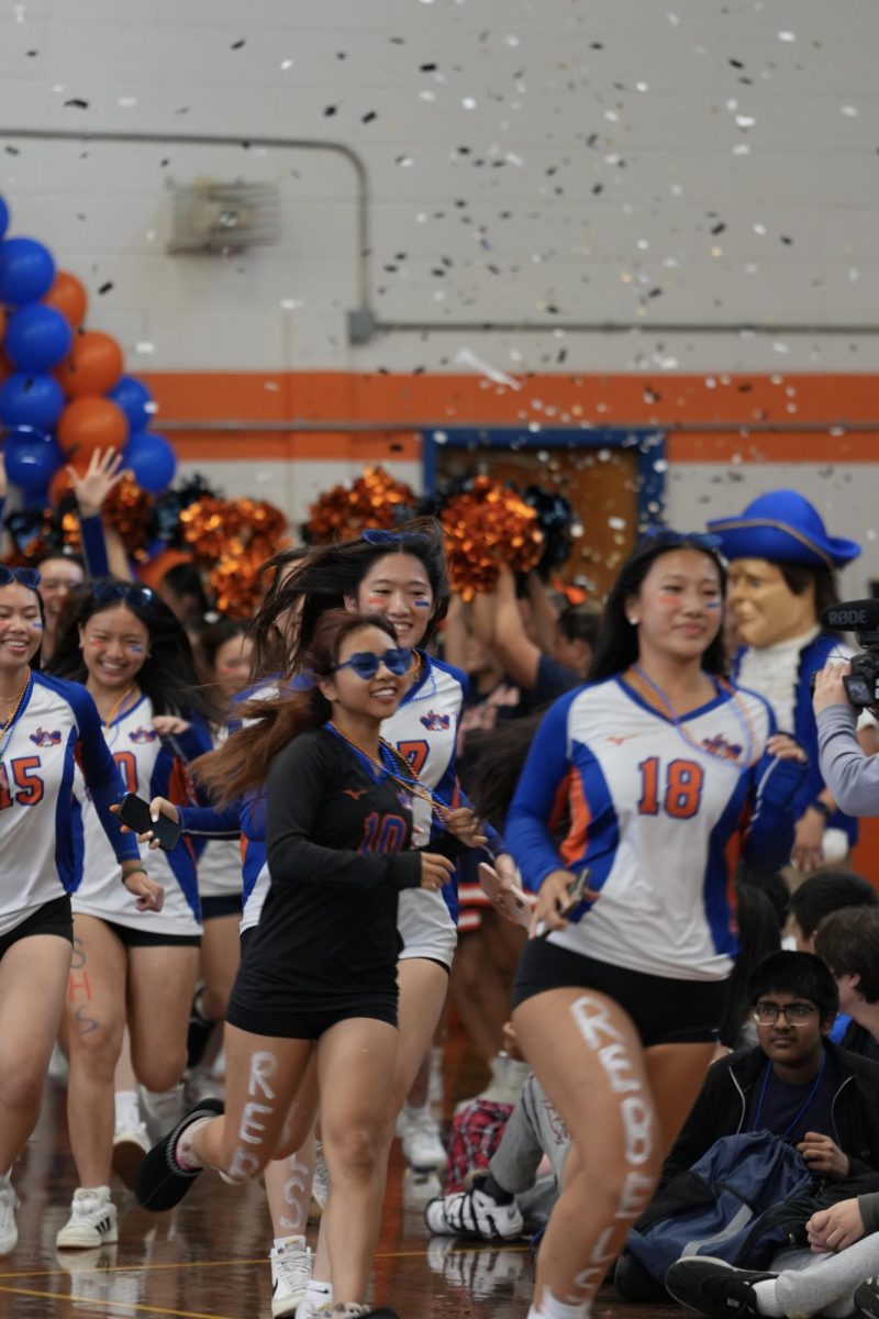 Girls Volleyball Sprints Through Confetti—Seniors Kelly Yu and Kary Wong lead the Girls Volleyball Varsity team as they enter the East Gym for Pep Rally. Wearing blue and orange colored necklaces and facepaints, the girls show rebel pride. 